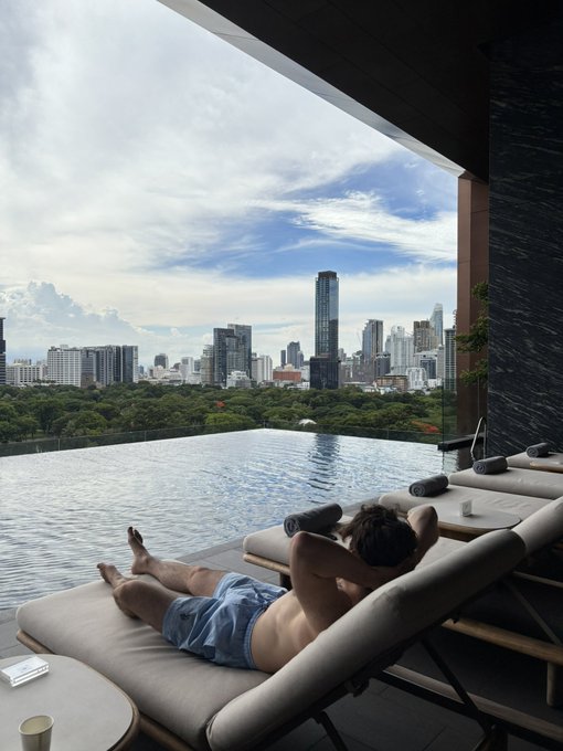 A man relaxes on a lounge chair by an infinity pool overlooking a city skyline, likely Singapore's.