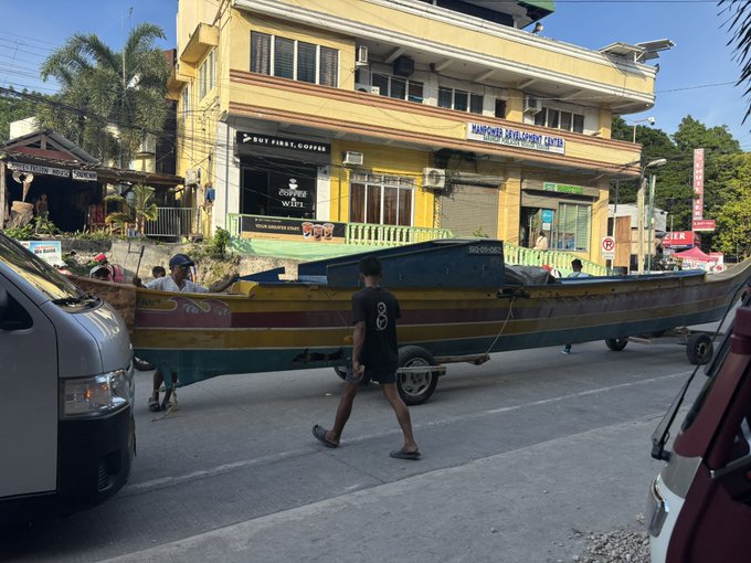 A man pulls a colorful wooden boat mounted on a trailer down a street past storefronts including "Mahinder Development Center" and "Buy First Coffee," playfully illustrating the post's tongue-in-cheek claim of having "bought a boat with Tezos."