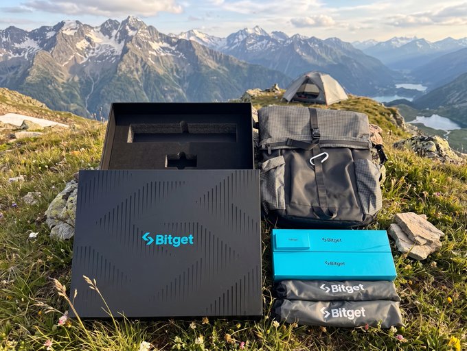 A Bitget anniversary gift box set, featuring the brand's logo and branded pouches, sits on a grassy mountainside next to a backpack and tent, showcasing the promotional package against a scenic alpine backdrop with snow-capped peaks.