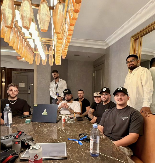 A group of about eight men gathered around a marble table in an upscale room with a decorative chandelier, featuring one man wearing a 'BIE' cap and a laptop displaying a sticker.