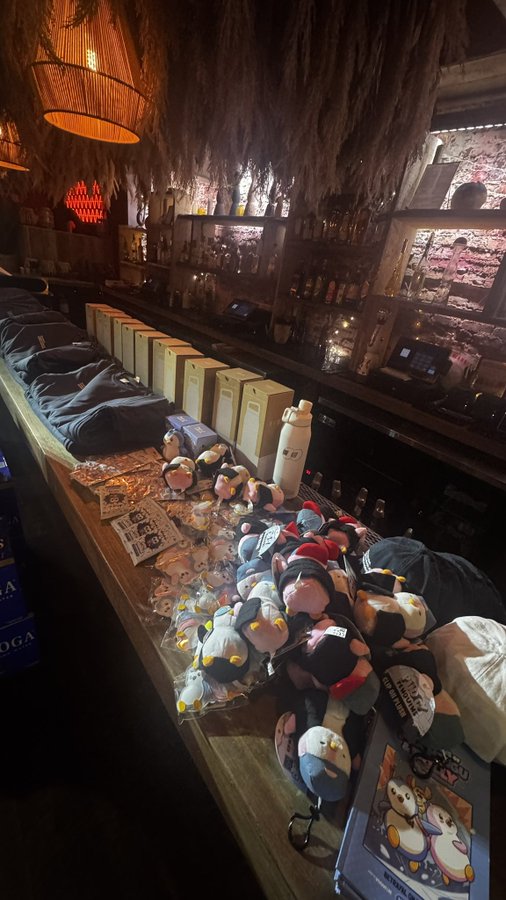 A dimly lit bar interior with a wooden counter displaying various penguin plush toys in plastic packaging and beige gift boxes along the bar surface.
