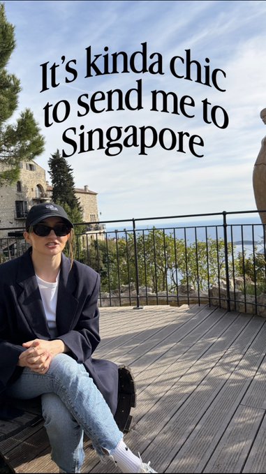 A person wearing a navy blazer, blue jeans, sunglasses, and a cap sits on a wooden bench with a scenic ocean view behind them, overlaid with the text "It's kinda chic to send me to Singapore".