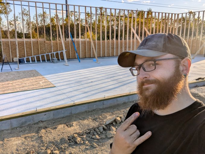A bearded man wearing glasses and a dark baseball cap points at himself while taking a selfie at a construction site, with wooden framing for a wall visible behind him.