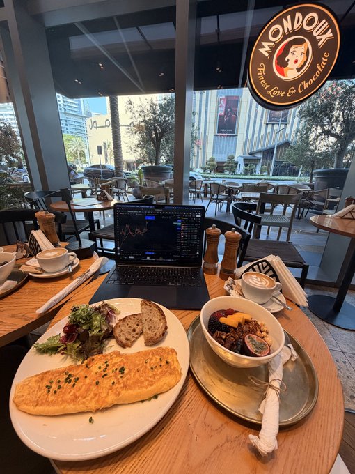 A meal at a Mondoux café (logo: 'Mondoux - Sweet Love & Chocolate') featuring an omelet, salad, bread, a fruit bowl, and coffee, with a laptop displaying financial charts open on the wooden table.