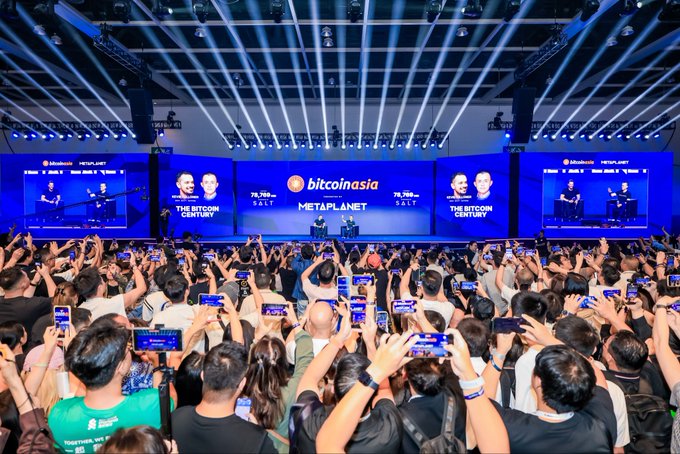 The image shows a massive indoor event at the Bitcoin Asia conference in Hong Kong, held on the "Nakamoto Stage" with the banner "bitcoin asia x METAPLANET" and "THE BITCOIN CENTURY" prominently displayed on large screens. A speaker stands at a podium on stage under dramatic blue and white spotlights beaming across the venue, addressing a crowd of over 3,000 attendees, many of whom are holding up smartphones to capture the moment. The scene visually matches the post's description of a packed Nakamoto stage with a "full house," capturing the energy of the chat with CZ, the founder of Binance.