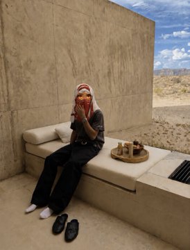 A young woman wearing a headscarf and casual loungewear sits barefoot on a built-in stone bench in a minimalist desert-style home, looking at her phone with food on a tray beside her. The barefoot, unassuming setting creates a stark contrast to the post's claim of having made $40 million from Trump, suggesting ironic detachment from immense wealth.