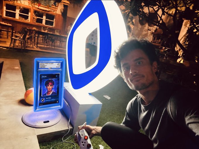 A man sits beside a glowing blue "Monad" logo, holding a small controller connected to a display showing his character card. The scene captures the festive atmosphere of "happy monad cards day," with the bright branding and personalized card suggesting a community celebration of digital collectibles.