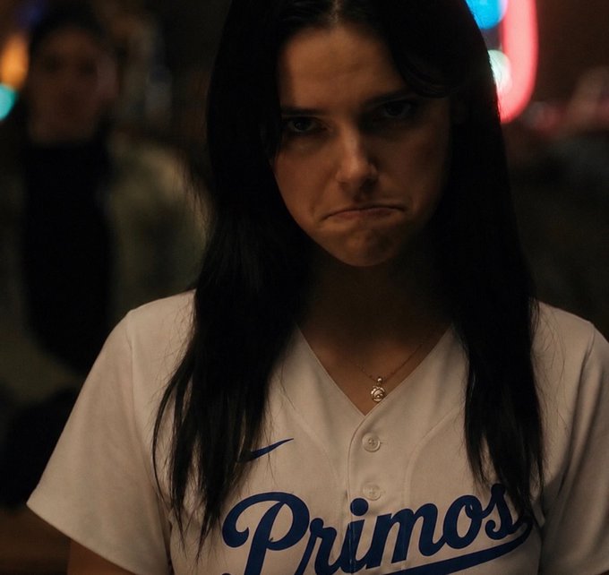 A young woman with dark hair wearing a white 'Primos' baseball jersey with a Nike logo looks displeased in a dimly lit setting.