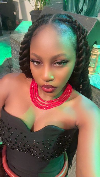 A selfie of a woman with braided hair wearing a black beaded top and a chunky pink/red beaded necklace in a green-lit setting.