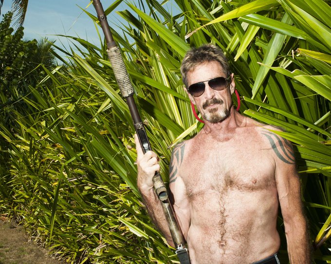 A shirtless older man with sunglasses, tribal tattoos on his shoulders, and a gray beard stands in front of lush green palm fronds, holding what appears to be a long rifle.