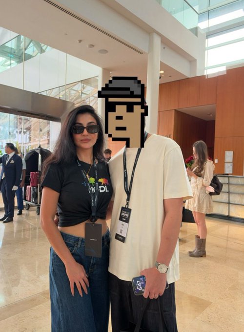 A woman with long dark hair and sunglasses stands next to another person whose face is obscured by a pixel art block, both wearing lanyards at what appears to be an event.