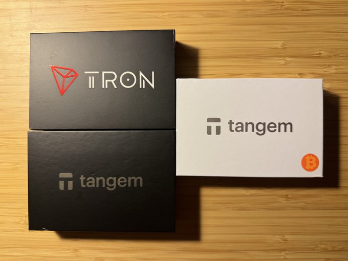 Three Tangem wallet card packages are displayed on a wooden surface, featuring two black "tangem" boxes and one with the "TRON" branding, alongside a white box showing the Tangem logo with an orange Bitcoin symbol. The image showcases the specific Tangem card sets being sold in the post, visually representing the three available bundles of 3 cards each offered at the discounted price of $30 apiece.