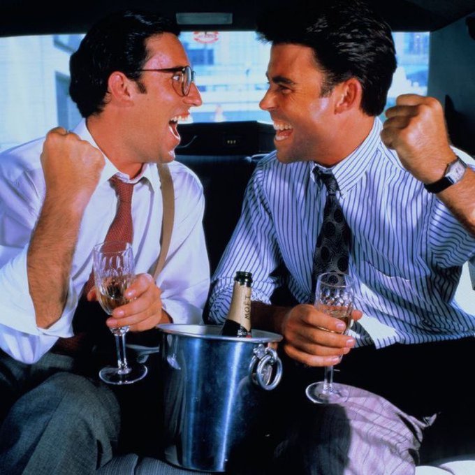 Two men in business attire sitting in the back of a car, laughing enthusiastically while holding champagne glasses with an ice bucket containing a Moët bottle between them.