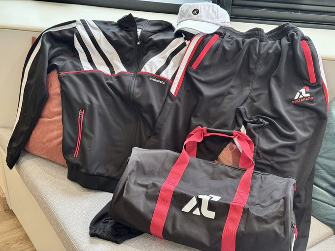 A collection of black and red 'everlastic' branded sportswear including a jacket, pants, cap, and duffel bag with 'AC' logo displayed on a couch.