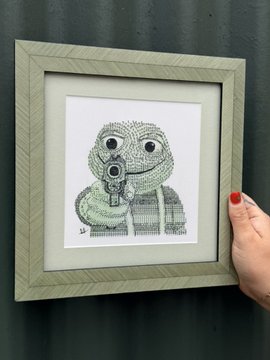 A hand with red nail polish holds a framed artwork depicting a small, bug-eyed character made entirely out of recycled computer keyboard keys.