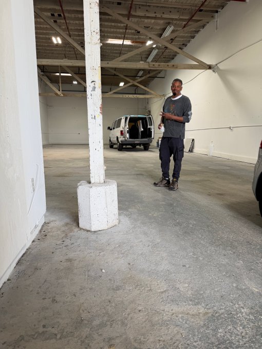 A man with a goatee, wearing a dark gray sweater and pants, stands in a large empty warehouse or industrial space with exposed ceiling beams and a white van parked in the background.