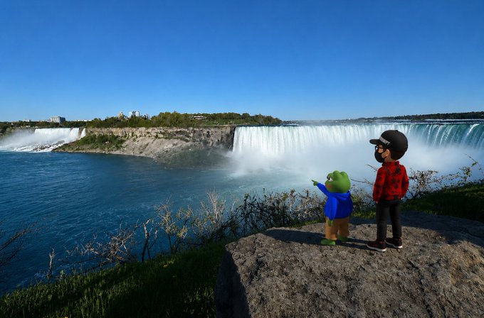 The image features two people standing on top of a rock, overlooking a waterfall. One person is wearing a blue jacket while the other has a red shirt. They are both looking at the waterfall with interest. The scene also includes a cityscape in the background, suggesting that they might be in an urban setting near the waterfall.