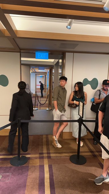 A group of attendees waits in line at the entrance of the Singapore Hyperliquid community event, with an illuminated "EXIT" sign visible above the doorway. The casual and stylish outfits of the participants reflect the lively, networking-focused atmosphere of the gathering.