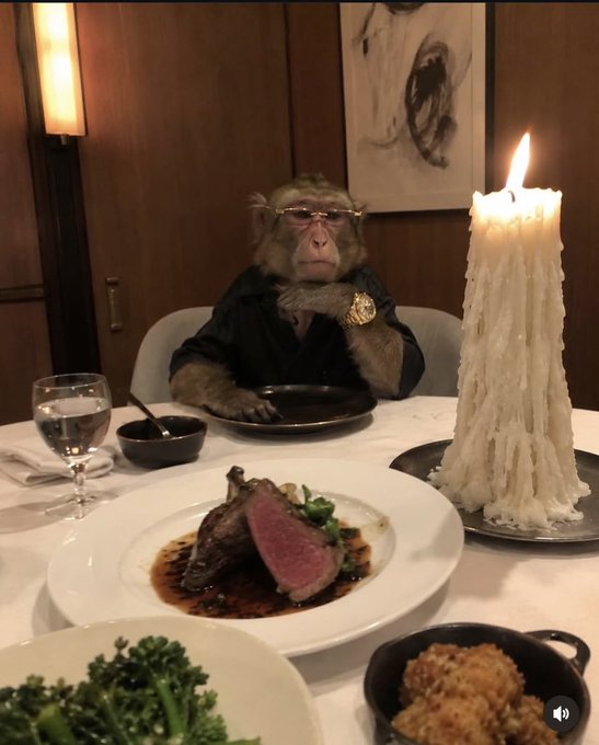 A monkey wearing a black shirt and glasses sits at a dining table with an empty plate, surrounded by dishes including a rare steak, a large lit candle, and drinks.