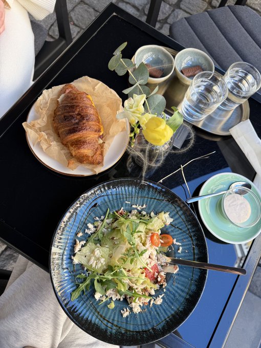 A black table set with a blue plate of salad topped with cheese, a flaky pastry on a white plate, a small vase with yellow flowers, and glasses of water.