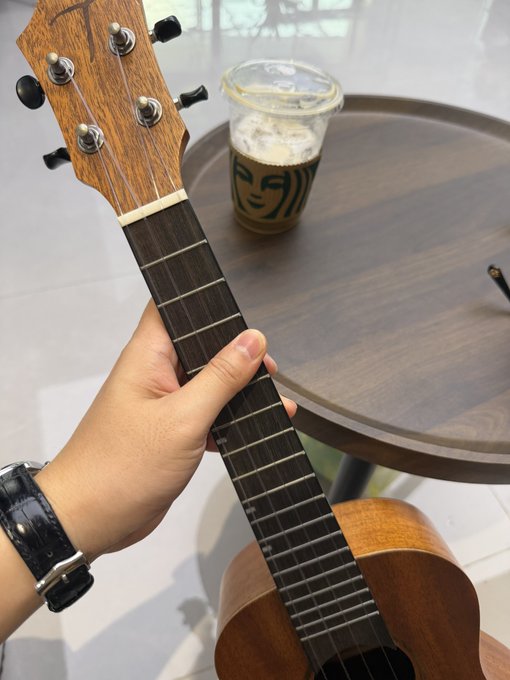 A hand wearing a black watch holds the neck of a wooden ukulele next to a wooden table with an iced coffee in a plastic cup.
