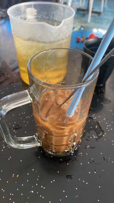 A photo of two beverages on a dark table - a glass mug containing a brown drink with straws and ice, and a plastic pitcher with a yellow liquid in the background.