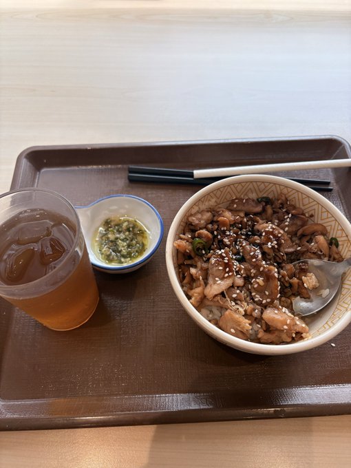 A meal on a brown tray featuring a bowl of rice topped with braised chicken, mushrooms, and sesame seeds, accompanied by a small dish of green dipping sauce and a glass of iced tea, with black chopsticks resting nearby.