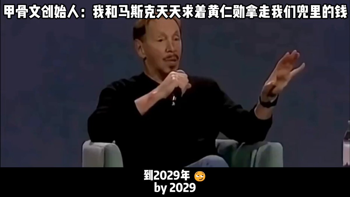 The image features a man sitting down, holding up his left hand with both hands in front of him as if he's talking or posing for a picture. The background has some text that reads "202009 by 2029", possibly referring to an article or video featuring the man. Additionally, there is another person visible in the image who appears to be talking or standing behind the main subject.