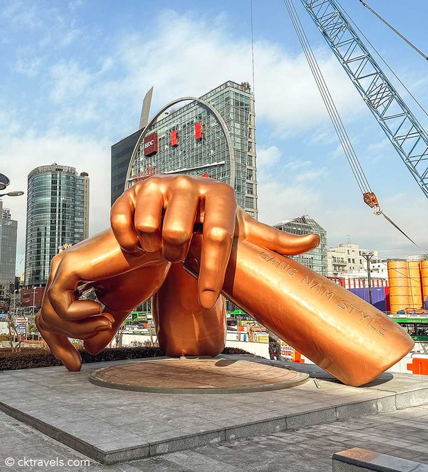 A massive copper-toned sculpture of two hands forming a finger heart gesture is displayed in an urban plaza, with "GIANG NAM STYLE" inscribed on one arm, set against a backdrop featuring an "HDC" branded building and construction cranes. The image ties to the Korea Blockchain Week post by showcasing a striking piece of public art in a Korean city, hinting at the vibrant, modern setting where the crypto and blockchain community is set to gather.