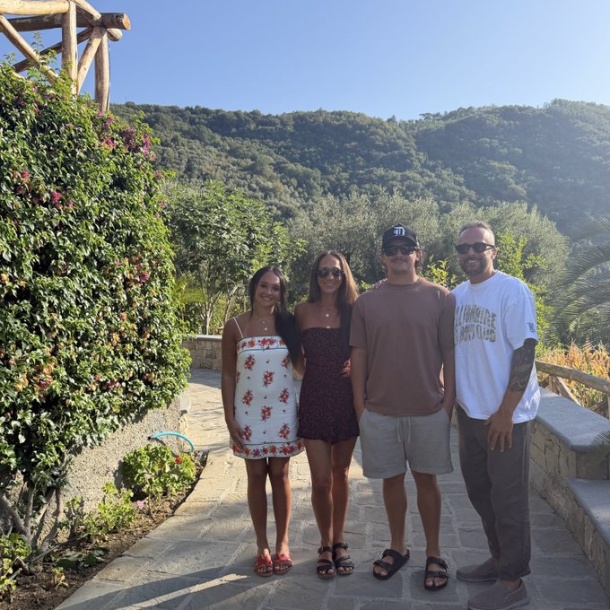 Four friends pose together on a sunlit stone pathway with lush green hills and vibrant bougainvillea in the background, one wearing a white "AT MARRAKECH" t-shirt, capturing a fresh travel moment that perfectly matches the "new beginning" vibes of the post.