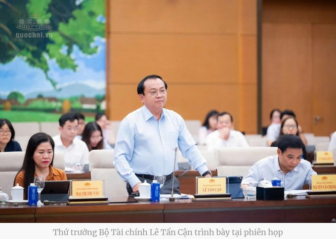A formal meeting setting where a man in a light blue shirt stands to speak; the caption identifies him as Deputy Minister of Finance Lê Tấn Cận, with the watermark 'quochoi.vn' visible.