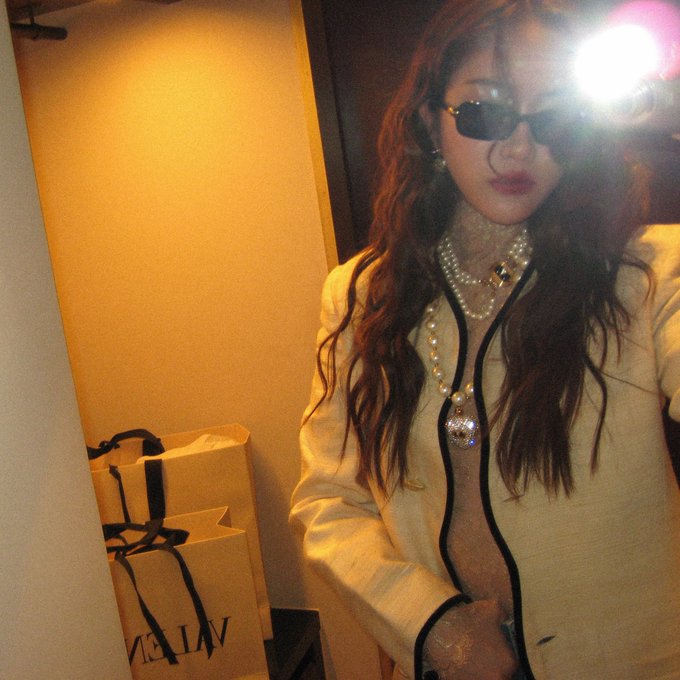 A woman with long wavy brown hair, sunglasses, and pearl necklaces poses indoors next to two shopping bags, with a bright camera flash visible in the corner.