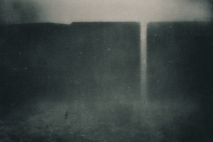 A grainy, dark, atmospheric black-and-white image depicts a massive, shadowy wall or dam structure with a bright vertical stream of water or light pouring down its center, while a tiny solitary figure stands in the misty foreground, dwarfed by the immense scale.