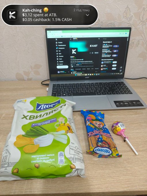 The image shows a laptop computer sitting on top of a wooden desk next to some snacks and candy bags. The snacks include chips and lollipops, while the candy includes a bag of Skittles. A person's name appears at the bottom of the photo, possibly indicating that they are the one who purchased these items or posted about them for others to see.