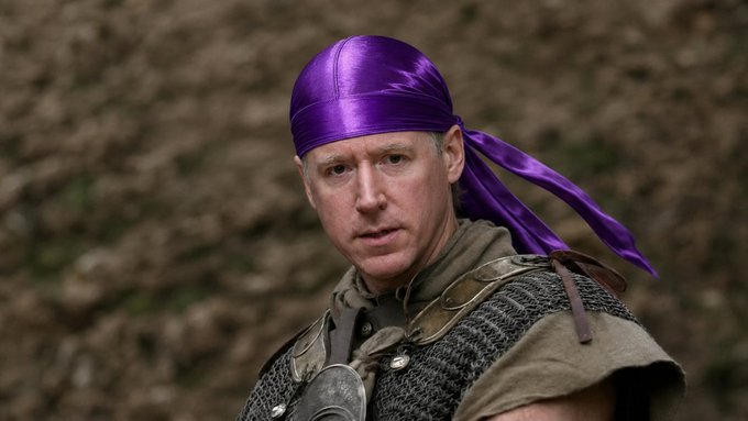A man in medieval-style armor and chainmail wears a purple satin headwrap with trailing fabric, photographed outdoors against a rocky hillside background.