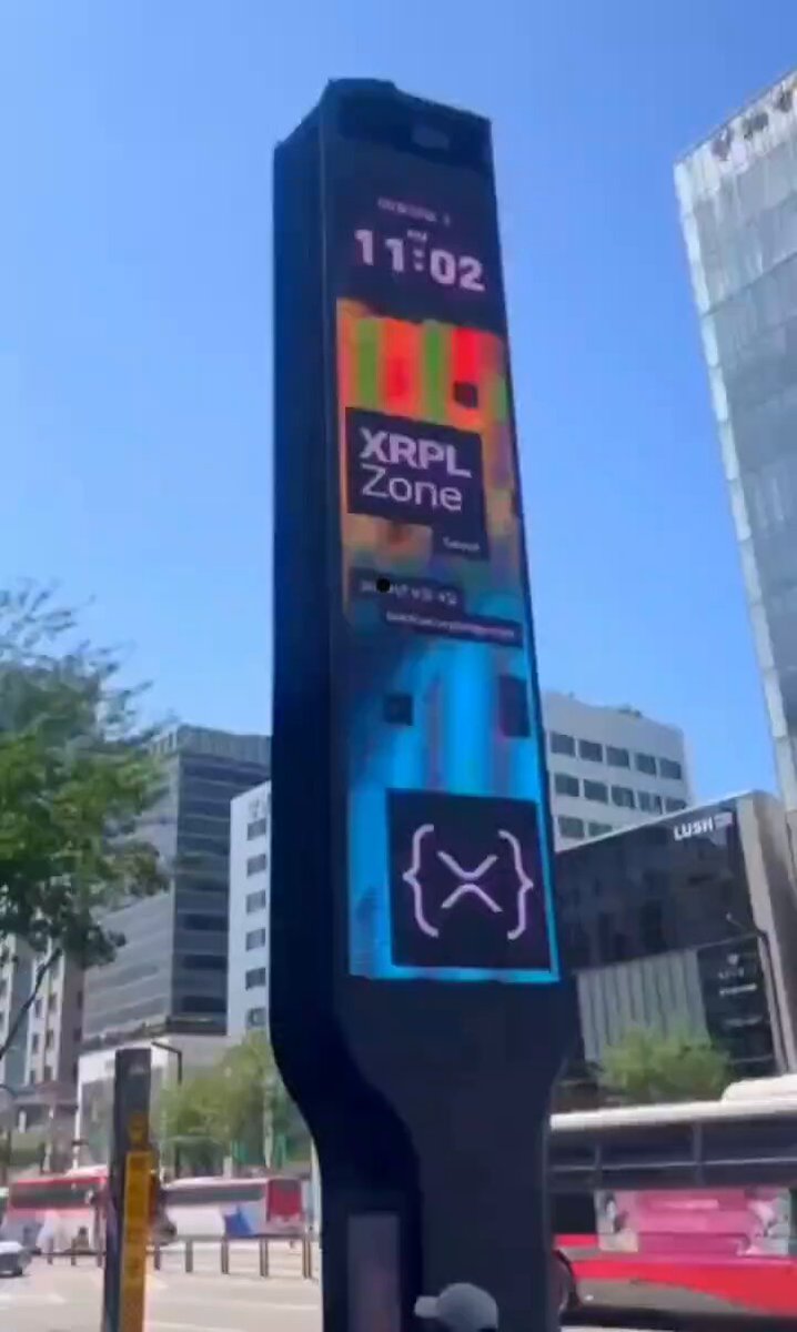 The image features a tall, black sign with colorful advertisements for XPPL and XRP. The sign is located at an intersection or bus stop, possibly indicating that XPPL is nearby. The text on the sign reads "WOW! THIS HUGE ADVERTISEMENT WAS SPOTTED IN SOUTH KOREA! XPPL IS ABOUT TO REVOLUTIONIZE THE WHOLE WORLD!". This suggests that XPPL might be a company with global reach and influence in the world of cryptocurrencies, as indicated by the colorful advertisements.