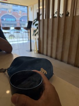 A first-person perspective photo of someone holding a dark ceramic mug while seated at a counter or table. A blue bag and a wooden slatted wall are visible in the background, with a window showing an outdoor building view.