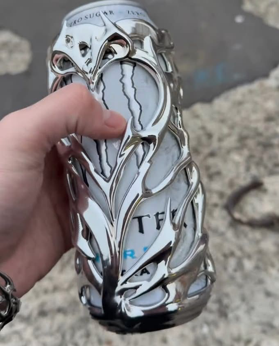 A hand holding a crushed silver 'TFL' (likely a Playboi Carti/The Filled Life) branded aluminum can with 'ZERO SUGAR' visible at the top, set against a concrete background.