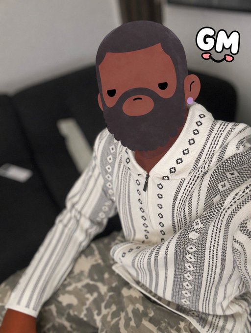 The user wants a concise caption for the image.
I need to look at the image and the provided text context.
- **Image Content:** A person is sitting on a couch. Their face is obscured by a cartoon avatar sticker (a man with a beard). They are wearing a patterned white and grey zip-up sweater and camouflage pants. There is a "GM" sticker in the top right corner with little pink cheeks.
- **Context Text:** "gm, Second day of the month and I’m craving more USDC."

**Connecting Image to Text:**
- The "GM" sticker in the top right corner directly relates to "gm" in the text (Good Morning).
- The text mentions "Second day of the month". This is likely just context for the date, not visually obvious, but the "GM" sticker confirms the "Good Morning" greeting.
- The text mentions "craving more USDC". This implies the person is talking about crypt