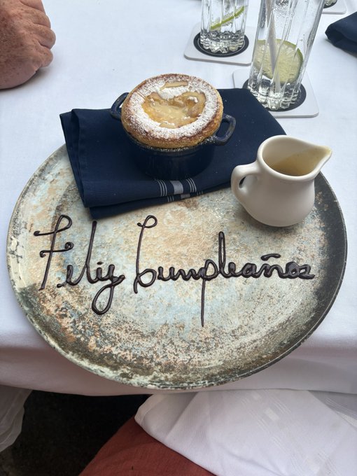 A dessert of "Feliz cumpleaños" (written in chocolate sauce on a rustic plate) is served at Monarca restaurant in El Salvador, celebrating a nurse's birthday lunch with what appears to be a powdered sugar-dusted pastry and a small pitcher of sauce.