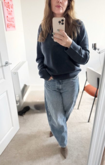 A woman takes a mirror selfie in a bathroom, wearing a dark blue sweater, light blue jeans, and brown boots.