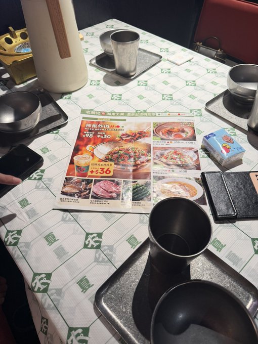 A restaurant table setting at a Hong Kong location of the mainland Chinese chain "农耕记" (Farm Bistro), featuring branded tablecloth, metal cups, bowls, and a menu advertising a 招牌鱼头 (signature fish head) for HK$98 plus a +$36 add-on. The image reflects the poster's complaint about being taken to eat this mainland chain in Hong Kong rather than local cuisine, highlighting the irony with promotional prices displayed on the menu.