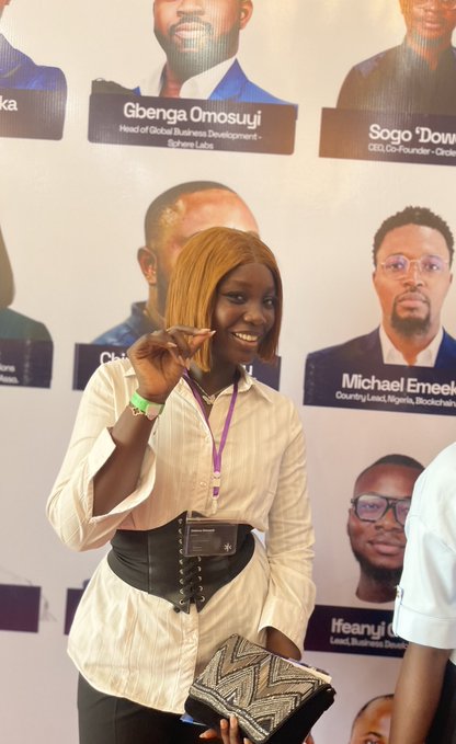 A smiling woman with short hair wearing a white blouse and lanyard, posing at what appears to be a tech event with a backdrop featuring names like 'Gbenga Omosuyi', 'Sogo', and 'Michael Emeka' with various professional titles.