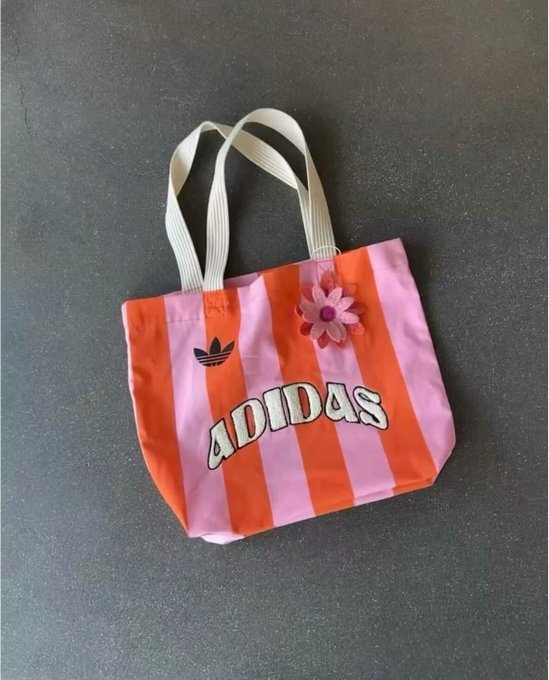 A pink and orange striped Adidas tote bag with white handles and a decorative flower, displayed on a dark surface.