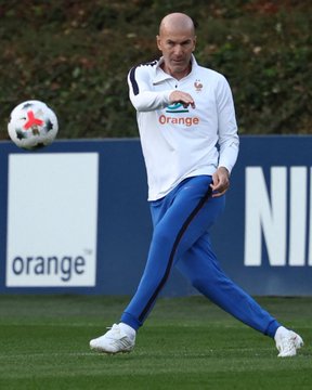 A bald man wearing a white France training jacket with the "Orange" sponsor logo and blue pants kicks a soccer ball on a training field, likely Zinedine Zidane in his role as France's national team coach, connecting to the upcoming France vs. Türkiye match mentioned in the post.