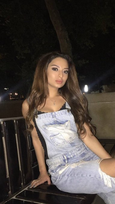 A young woman with long, wavy highlighted hair sits on a bench at night, wearing light blue denim overalls with distressed details over a black top, and a small cross necklace.