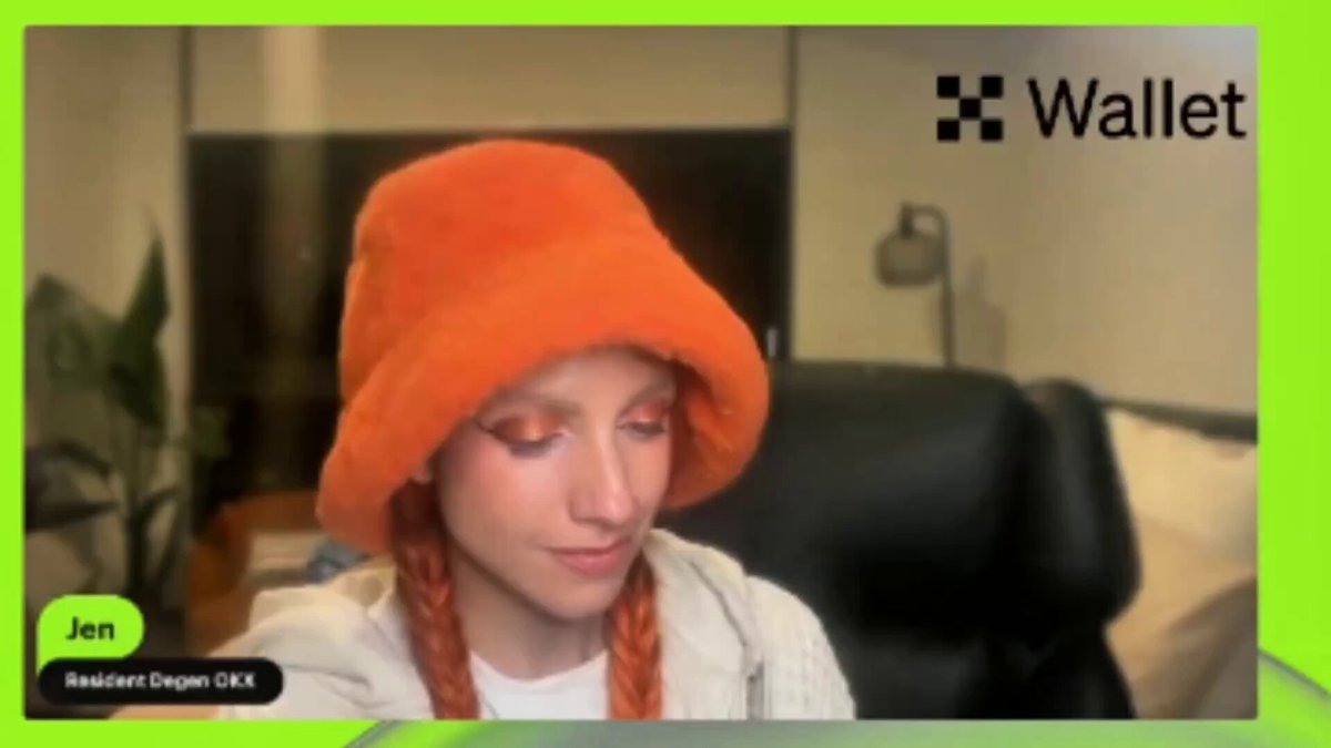 The image features an orange hat with red hair, which appears to be worn by a woman sitting at a desk or table. The background of the image shows a green screen displaying a message about a new game called "Emblem". This suggests that the woman might have been involved in creating or testing this game.