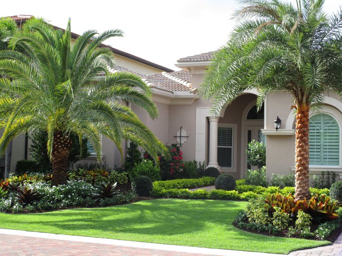 A well-manicured suburban home with a lush green lawn, tropical landscaping including large palm trees, and a Mediterranean-style exterior with terracotta roof tiles.