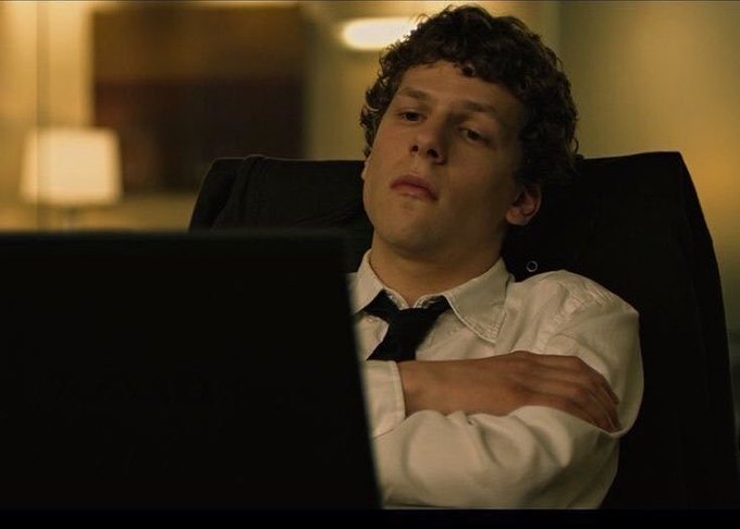 A young man with curly hair wearing a white shirt and loose black tie sits in a dark office chair in front of a laptop.