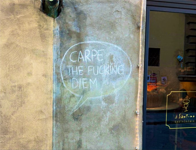 Chalk writing on a wall outside a shop reads 'CARPE THE FUCKING DIEM' inside a speech bubble, next to a window displaying 'il SogTino GASTRONOMIA'.